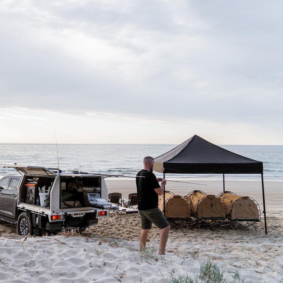 Person standing next to a van and beach setup with a canopy on a sandy beach.